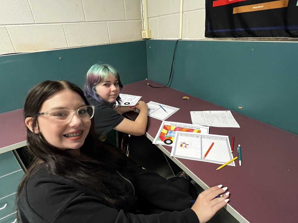Two girls seated at a desk, one wearing glasses, both smiling, working on papers with colorful markers.
