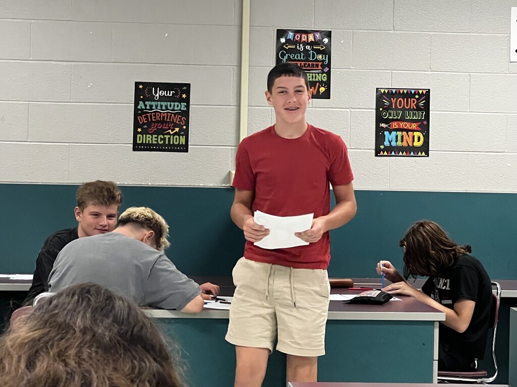 A man stands before a group of students in a classroom, holding a piece of paper. Behind him, motivational posters adorn the walls.