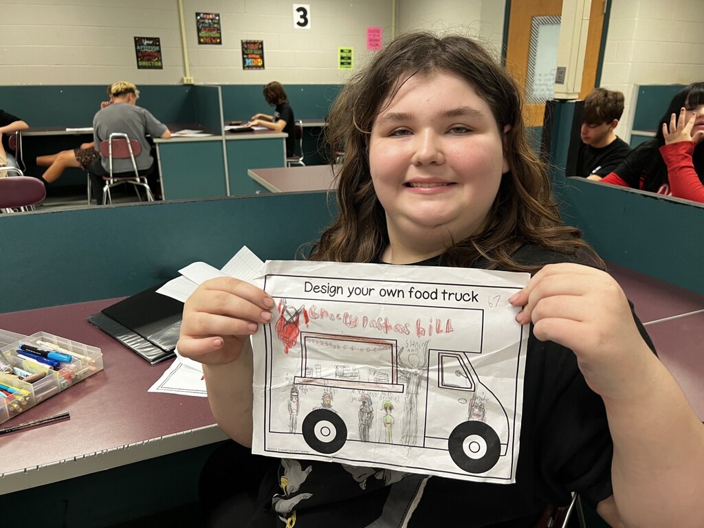 A person holds a drawing of a food truck with people inside. A desk and a few people are in the background.