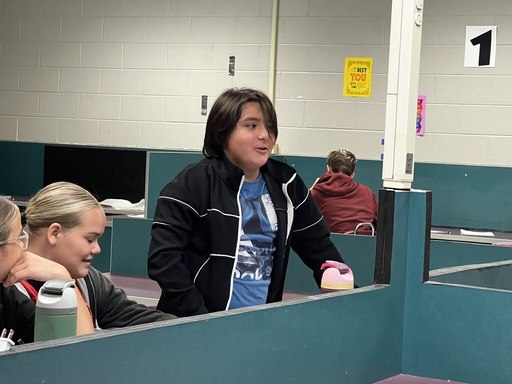 Three individuals sit in partitioned cubicles. One wears a blue shirt, another a black jacket, and the third wears a red hoodie.
