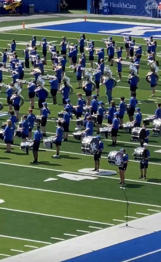 A marching band practices on a football field. Blue uniforms, brass instruments, and drums are visible. The field has blue markings.