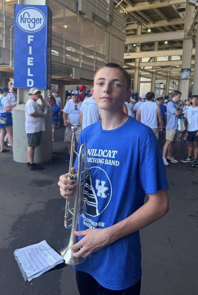 A young man in a blue Wildcats marching band shirt stands holding a trumpet while others walk behind him.