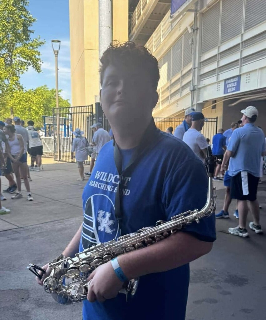 A young person in a blue shirt holds a saxophone. In the background, people walk on a sidewalk.