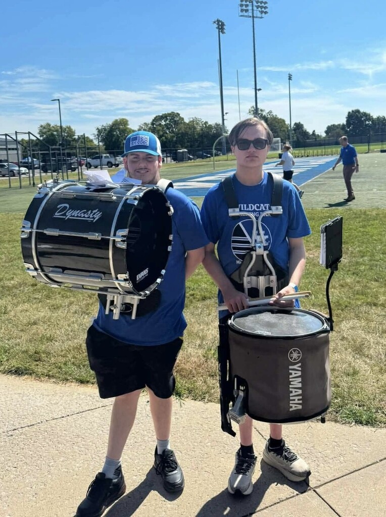 Two young men, both wearing blue shirts, stand on a path with drums. One holds a black drum, the other a Yamaha drum. Behind them, a grassy area and distant lights.