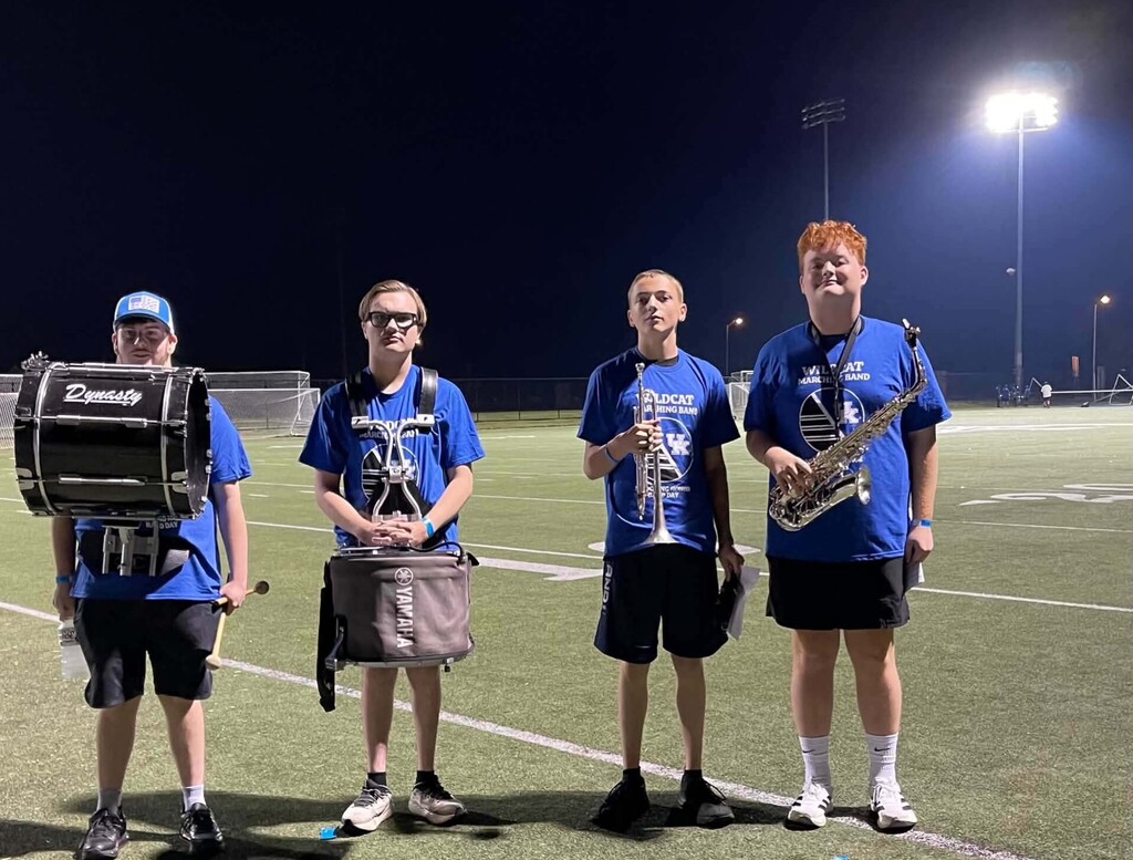 Four young individuals in blue shirts and shorts, holding musical instruments, pose on a field at night.