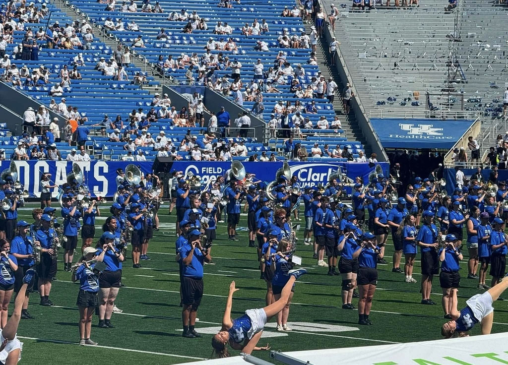 A football field with blue bleachers and people. A marching band stands in formation and two performers execute stunts.