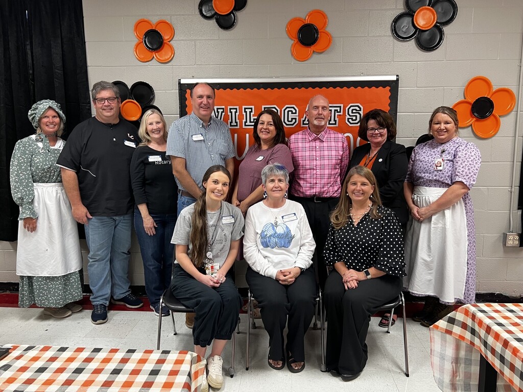 Group of people in a room with Halloween decorations, a sign reading "Milocats" and tables covered with orange and black tablecloths.
