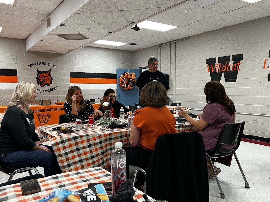A group of adults sits around tables in a room with a white wall, orange and black decorations, and a black chair.