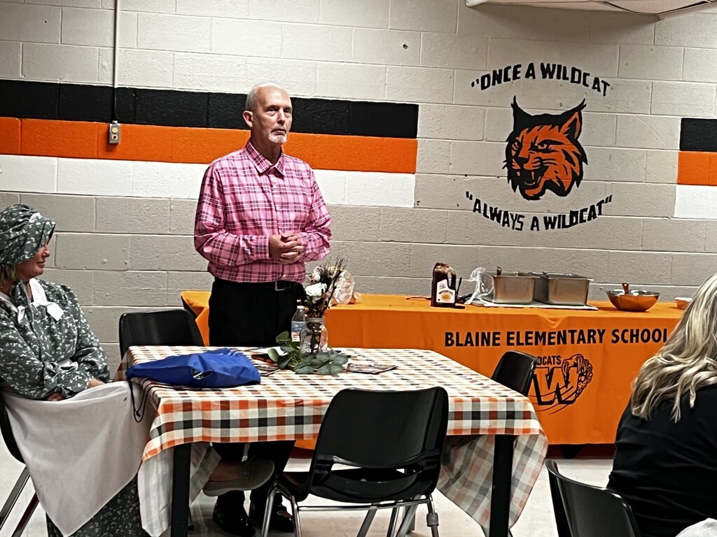 A man in a pink shirt stands before a table in a room with checkered tablecloth. Behind him, a banner reads "Once a Wildcat, always a Wildcat."
