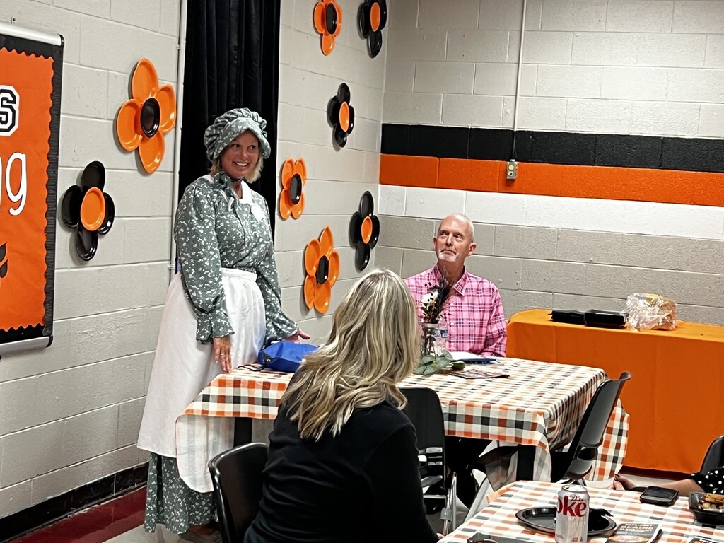 A woman in a bonnet and dress stands by a table. Two people sit at the table. A woman has a plaid tablecloth.