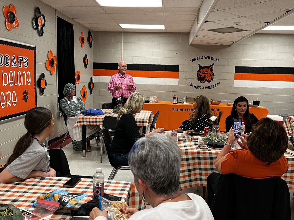 A room with tables and chairs, an orange wall, and a man in a pink shirt stands at a table.