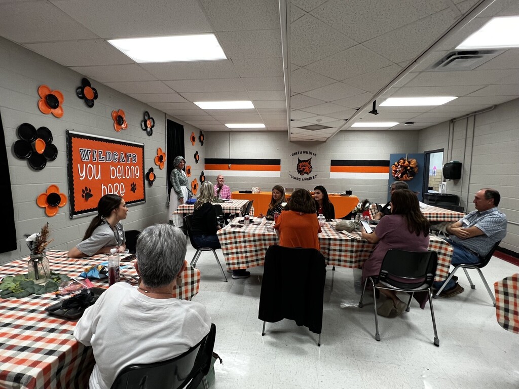 Room with tables and chairs. People seated around tables with tablecloths. Walls adorned with orange flowers and banners.
