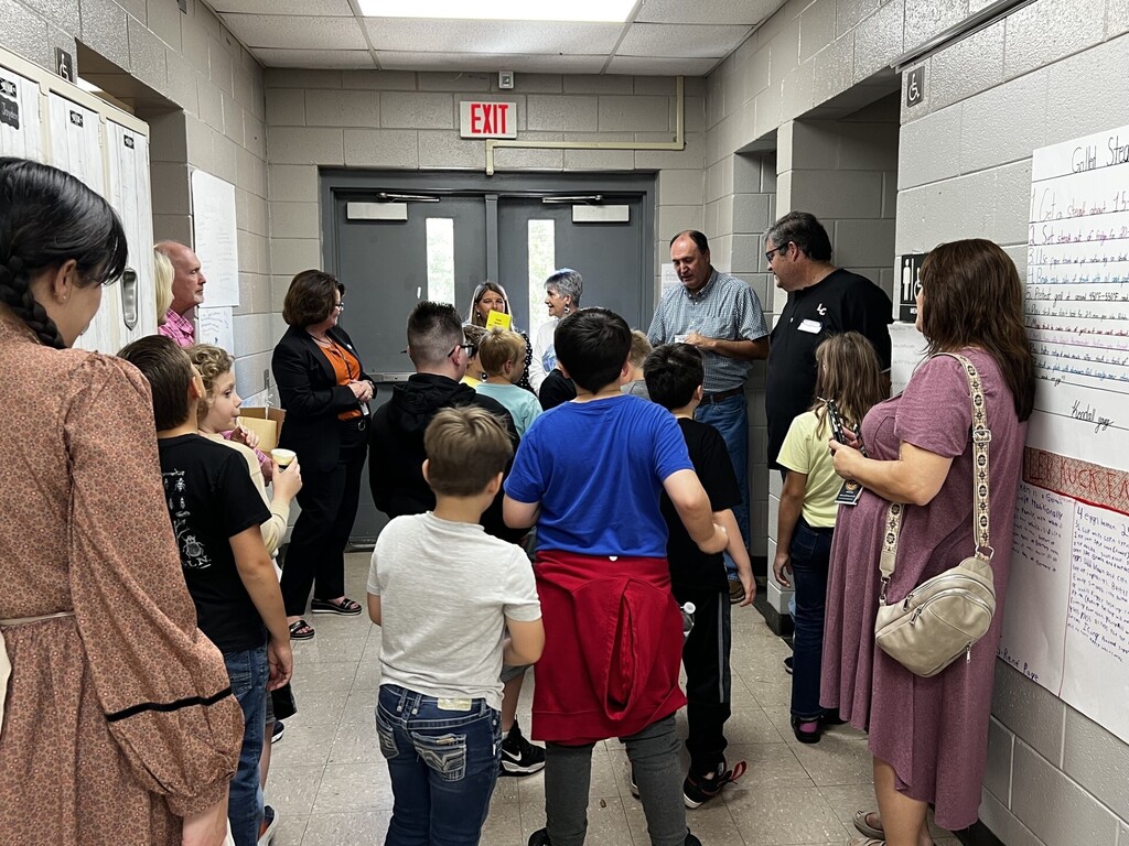 Several children and adults in a corridor, some holding papers. Bright white walls and doors.