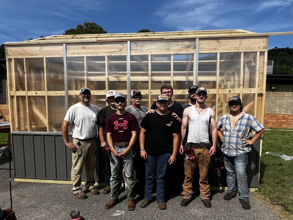 Group of people in hats and work attire stand in front of an unfinished wooden building.