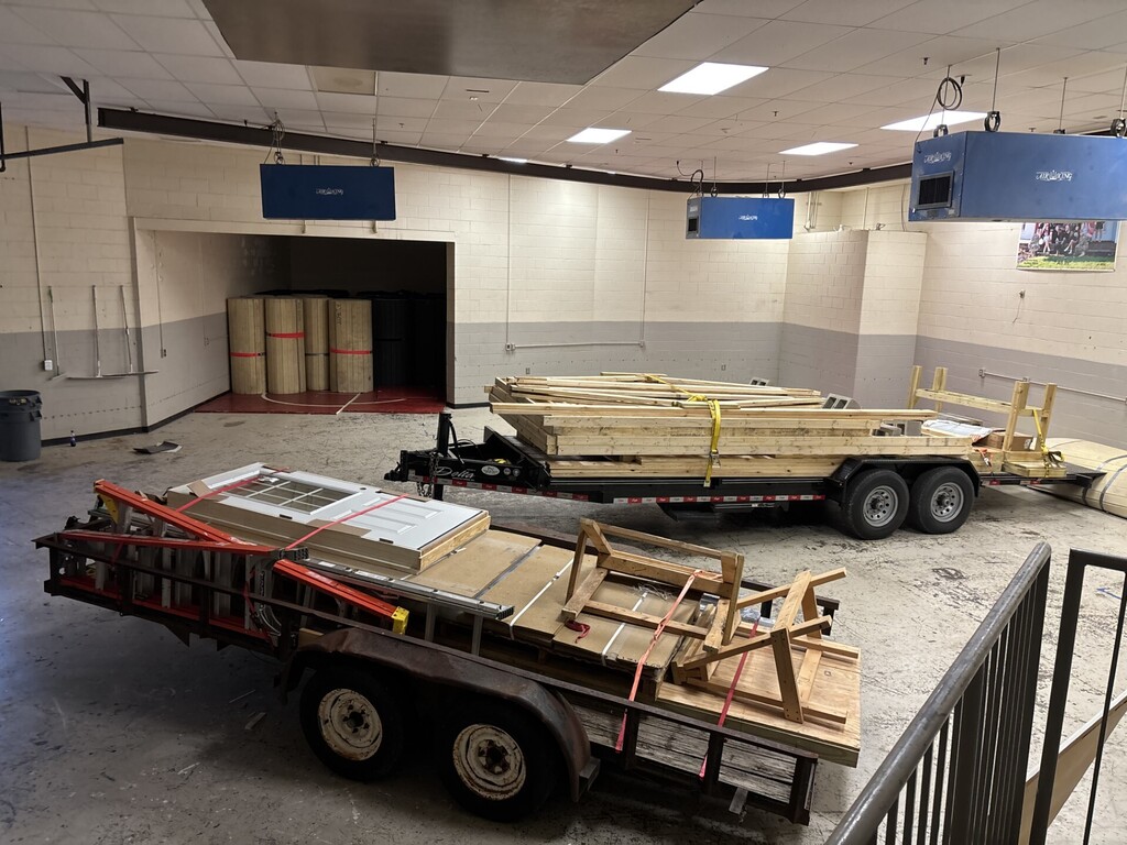 Two flatbed trailers with lumber and tools sit in a room with a metal railing and two blue signs.