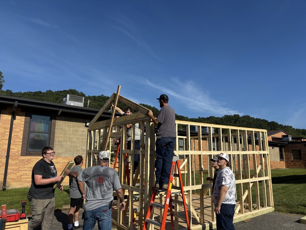 People work on a wooden structure outside a building with a sloped roof. Some on ladders.