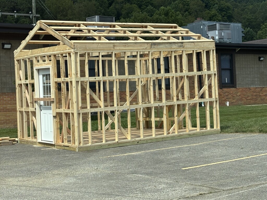 A wooden structure under construction, resembling a small house, sits in a parking lot. It has a roof and a door.