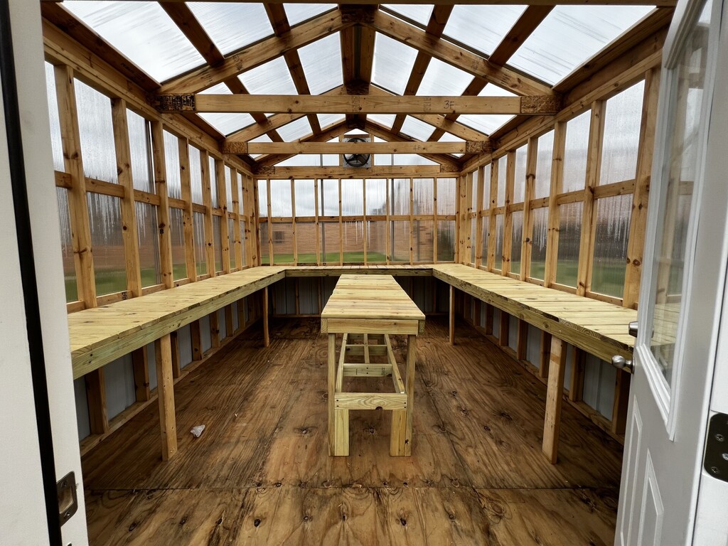 Greenhouse interior with wooden benches, a wooden table, and a white door open to a field.