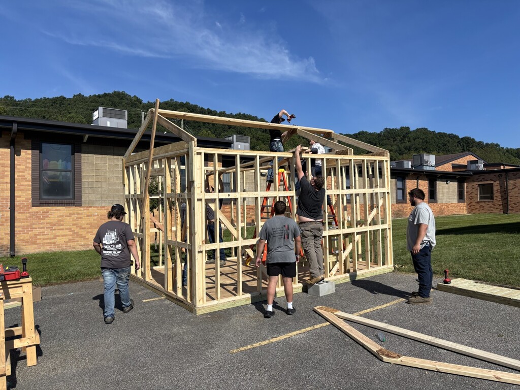 People constructing a wooden structure in a parking lot. A brick building and grassy hill in the background.