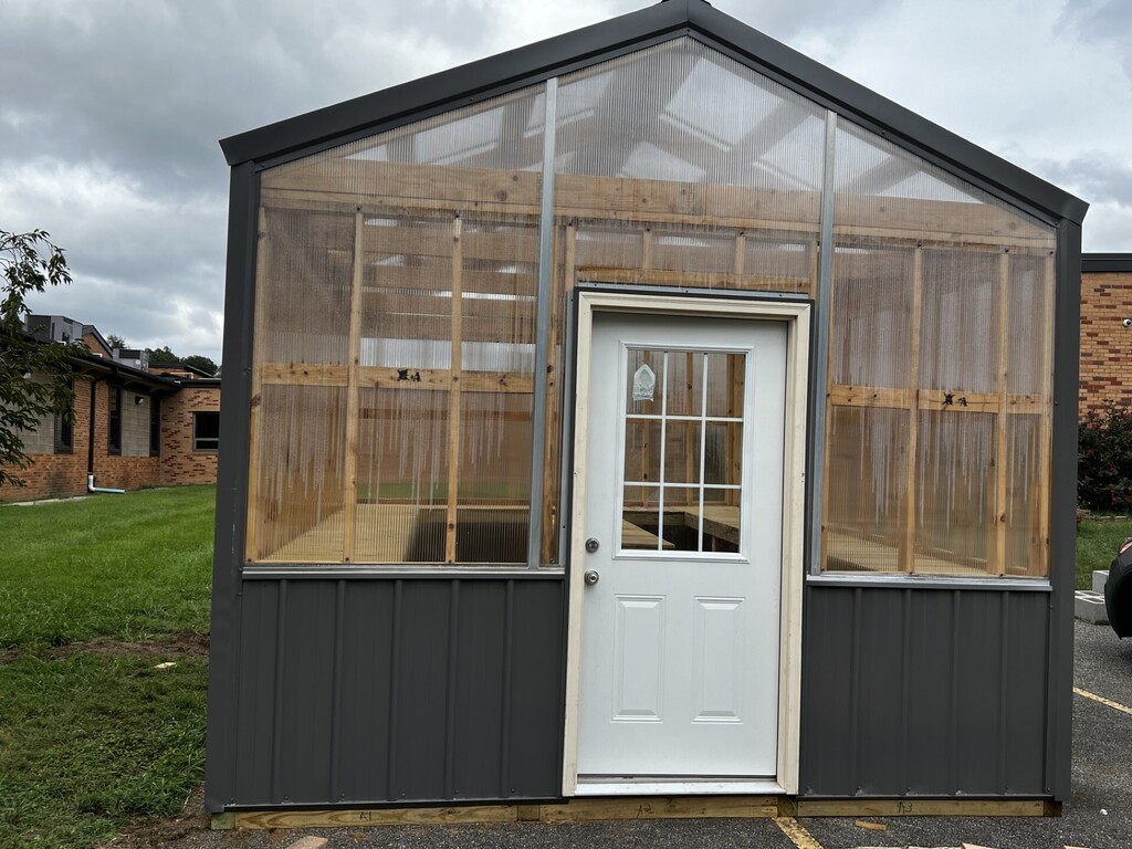 Greenhouse under construction with mesh windows and a white door. Grey exterior and green grass in the foreground.