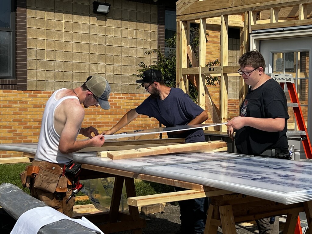Three young men work on a workbench outside, wearing hats, glasses, and one with a belt bag.