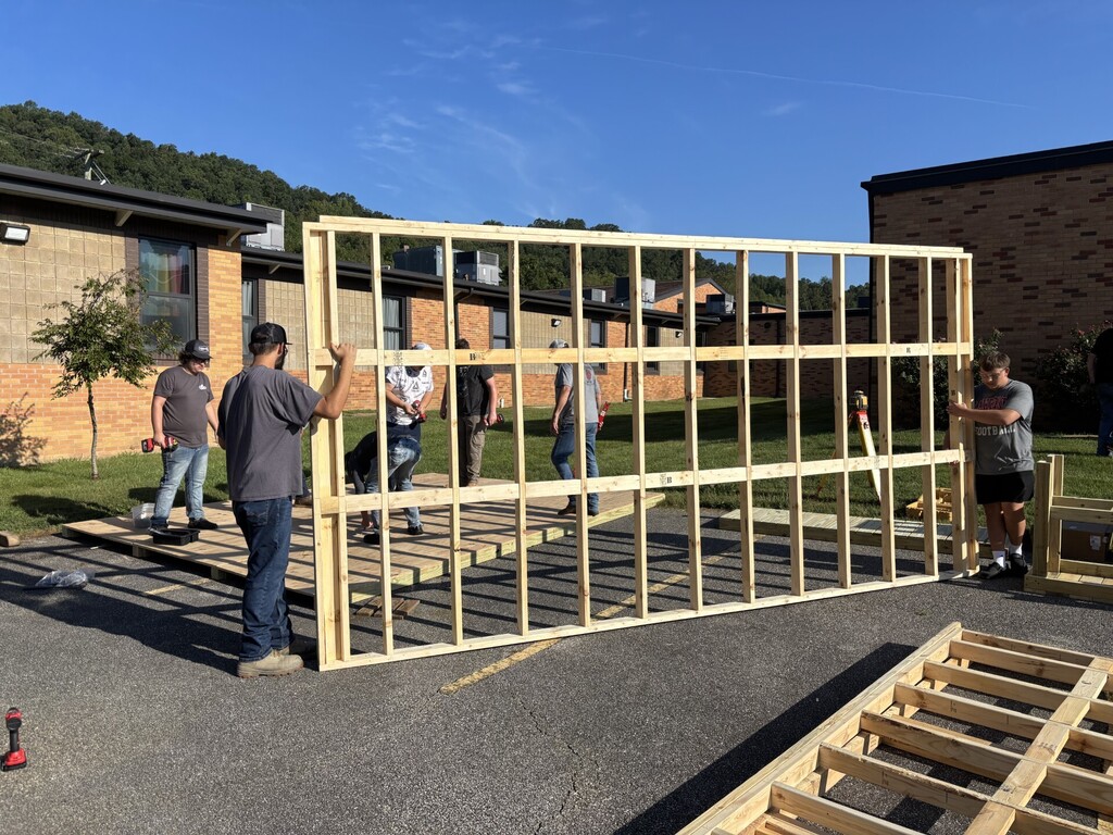 People work on wooden structure outside school. Some hold frame, others stand nearby. Background includes school building and grassy area.