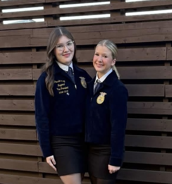 Two girls in matching navy jackets and ties stand close to each other in front of a wooden wall.