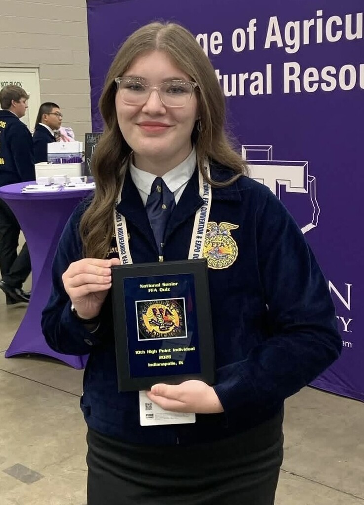A smiling young woman wearing glasses and a school uniform holds a framed award in her hands.