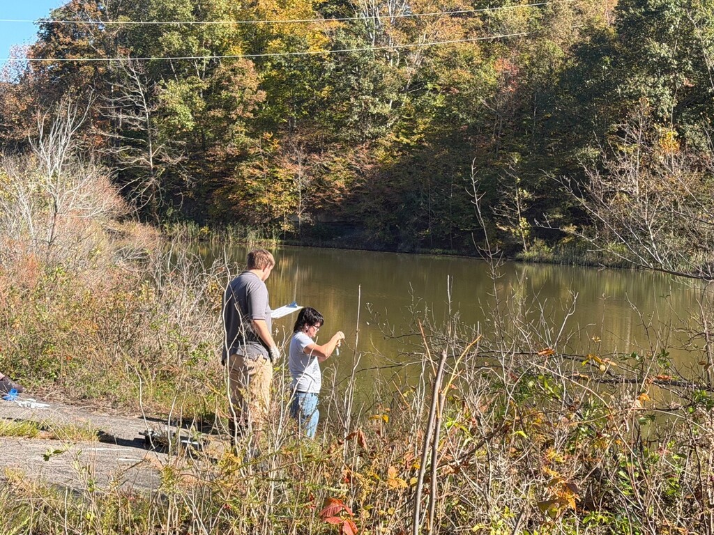 People standing by a pond on the edge of a wooded area, one holding a small container.