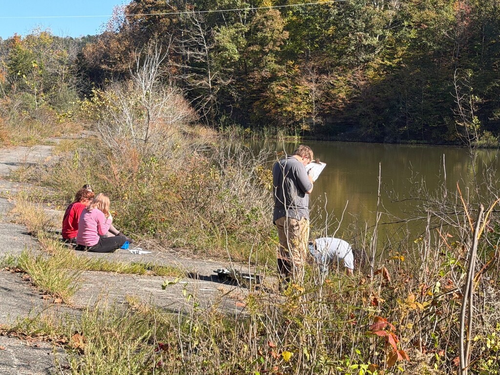 People sit on path near water with one man standing and reading. Trees line the shore.