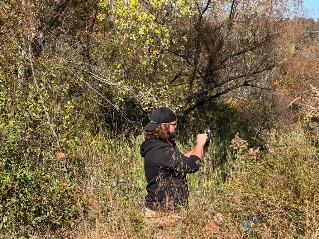 Man with beard in a wooded area, wearing a black hat and jacket, holding a camera.