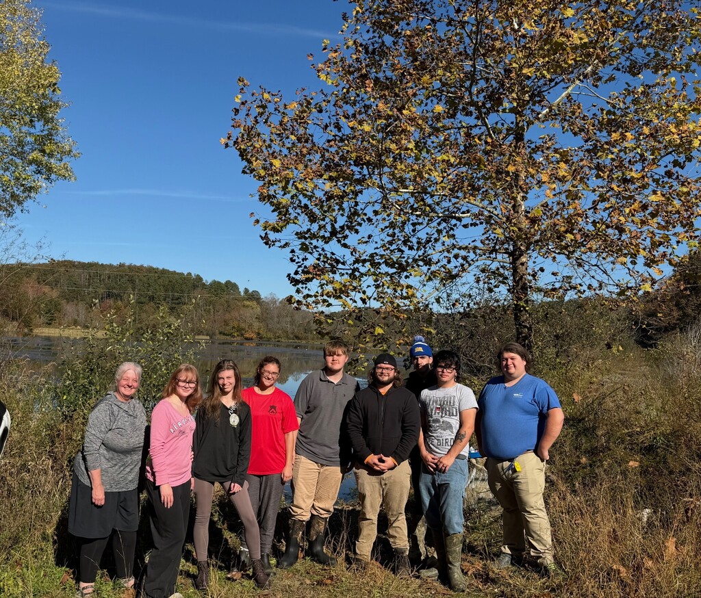 Nine people in casual clothing and boots pose in a field with a river in the background.