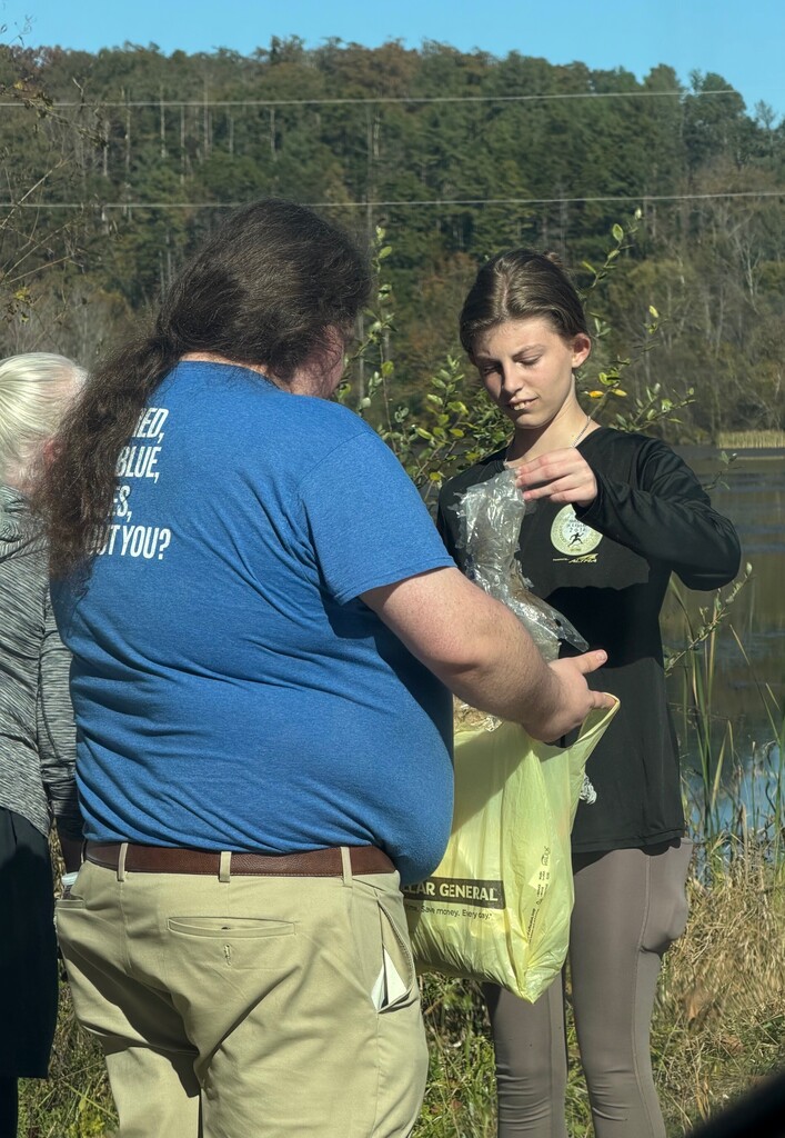 Two people near water, one holding a yellow bag, looking at a wrapped object. Background features trees and power lines.