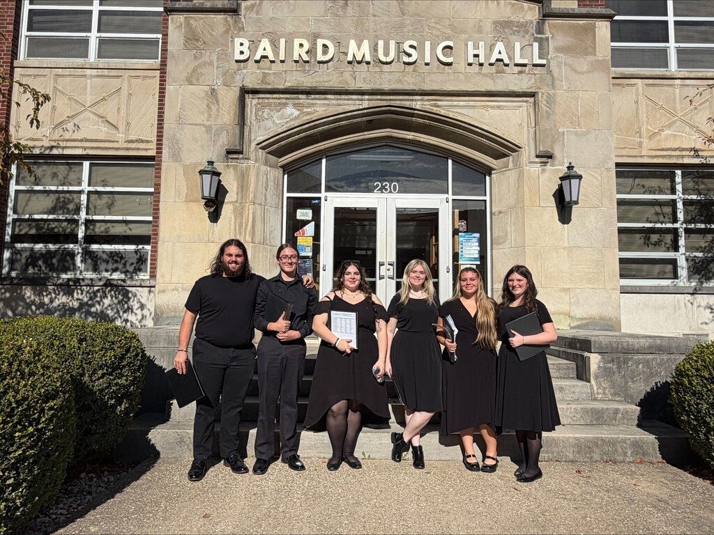 Group of people in black dresses standing on steps in front of a brick building with sign.