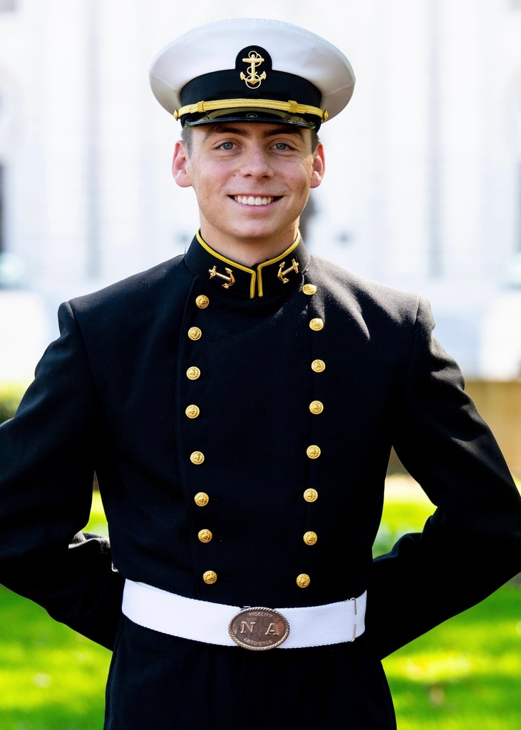 Matthew Aguilar in a navy uniform stands with a smile. The uniform includes a white hat and gold buttons.