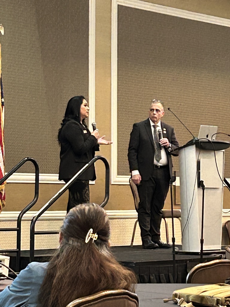 Two people stand on a stage with a podium, one holding a microphone. A person sits in the audience, facing the stage.