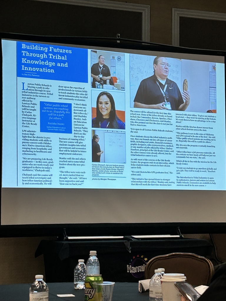 Presentation screen shows text and images of people. A table with cups and bottles is in the foreground.