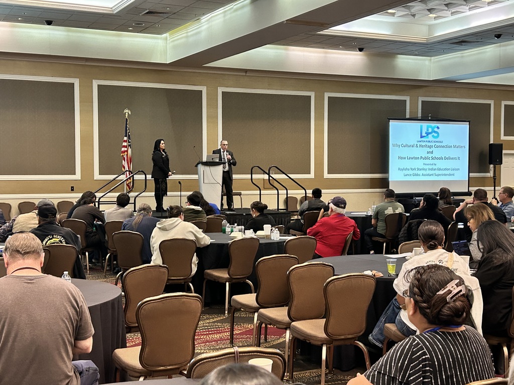 A conference room with a projector screen. People sit at tables and listen to speakers on stage.