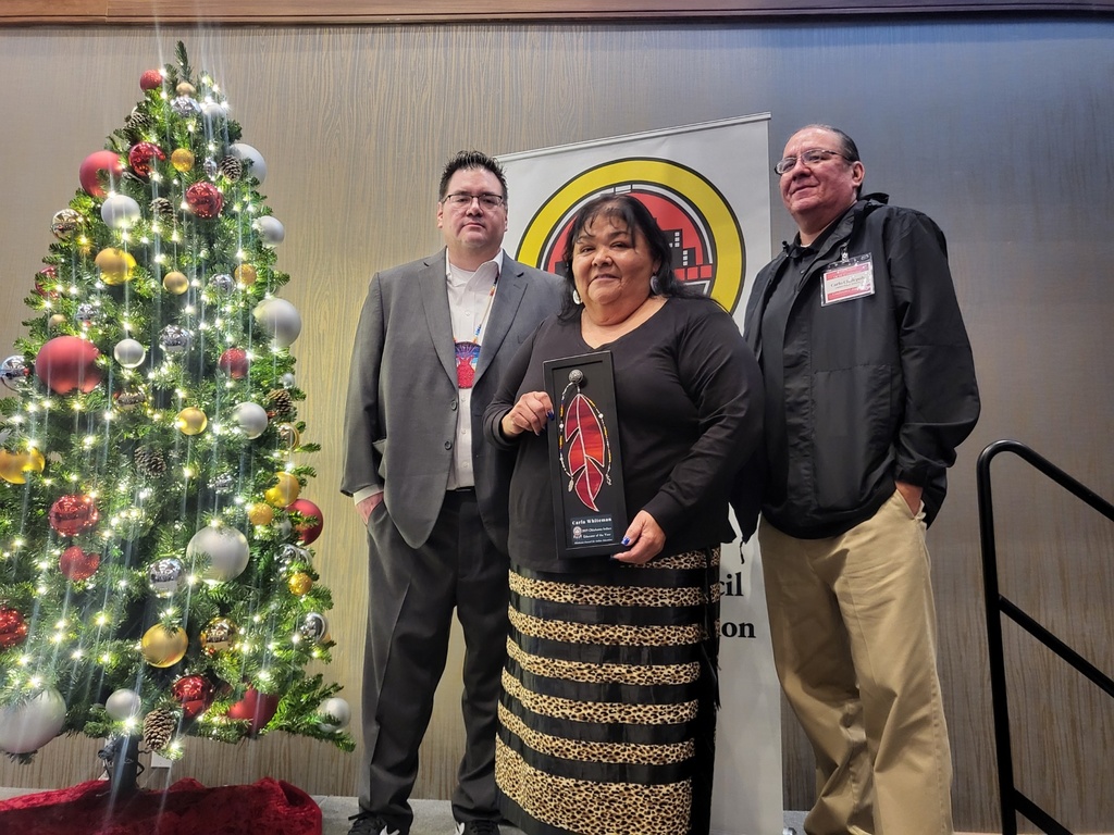Three people stand near a decorated Christmas tree and a sign. A woman holds a trophy, while two men stand behind her.