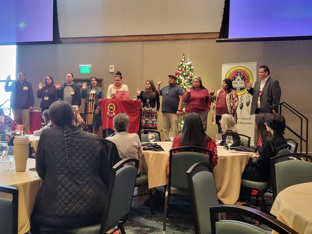A room with tables, chairs, and a Christmas tree. A group of people stand near a podium with a red cloth.