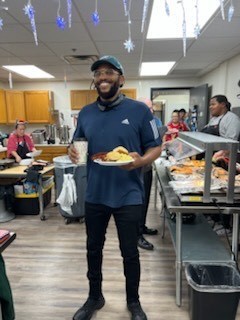 A person holding a plate of food in a cafeteria. Trays of food, people, and wooden cabinets are in the background.