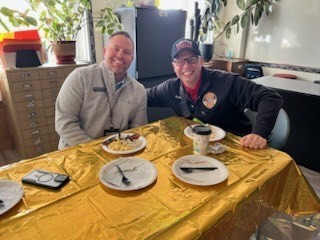 Two men in jackets and caps sitting at a table with plates of food and drinks.