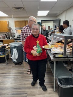 A woman in a Christmas sweater holds a plate of food in a cafeteria. People gather behind her.
