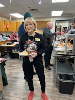 A woman with short blonde hair holds a plate of food in a kitchen with wooden cabinets.