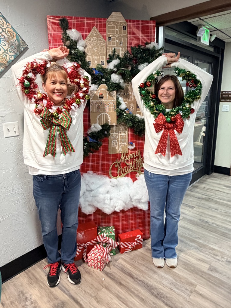 Two women stand near a Christmas wreath decoration, both wearing white sweaters and wreaths around their necks.