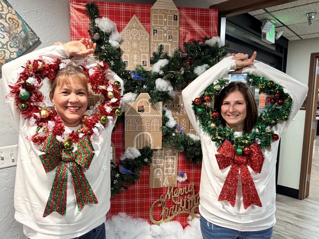 Two women wear Christmas sweater decorations, posing with hands raised. Behind them, a gingerbread house and Christmas wreaths are displayed.