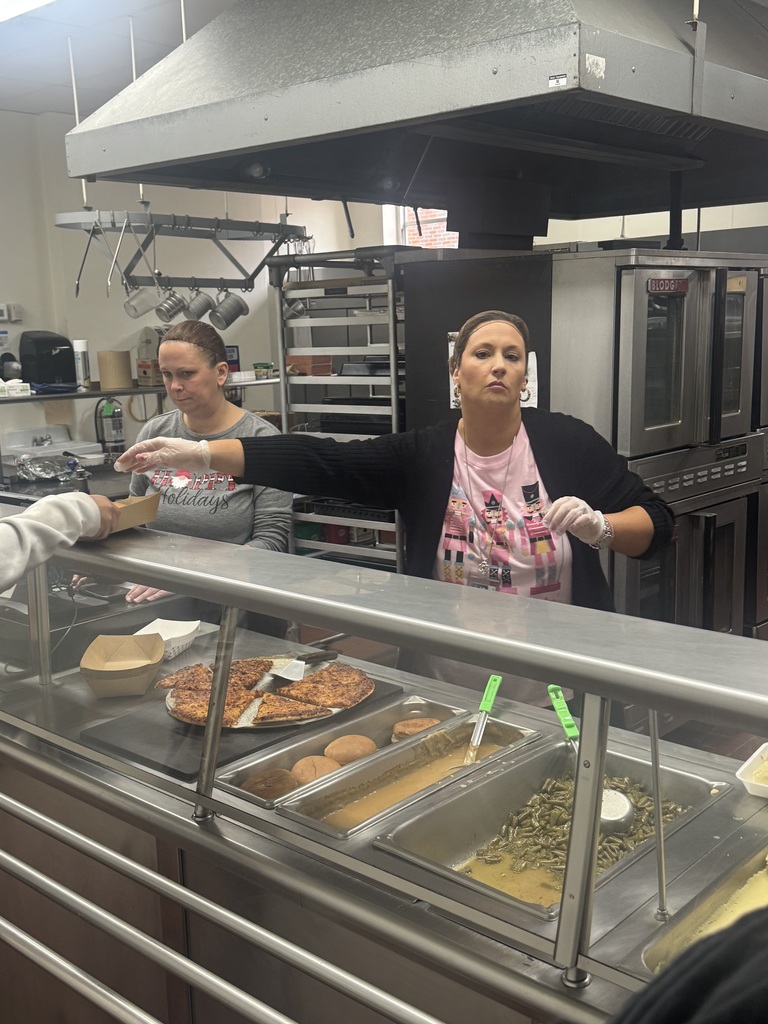 Two women in a kitchen area. One woman serves food from a tray, while the other takes food from a serving window.