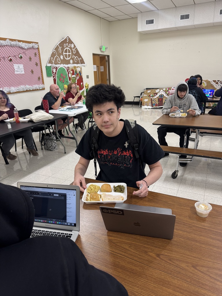 A person holding a tray of food stands at a table in a classroom with others seated.