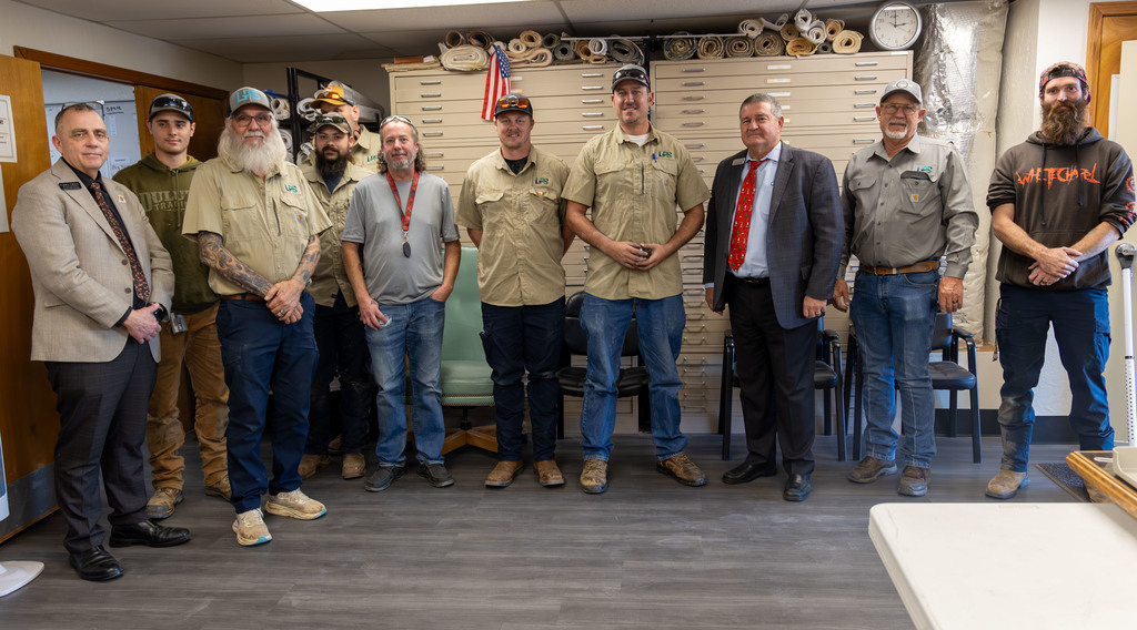 A group of people wearing hats and some with beards, dressed in casual work attire, stand in a room.