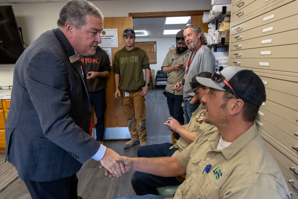 A man in a suit shakes hands with another man in a tan shirt. Behind them, several men in casual clothing stand around.
